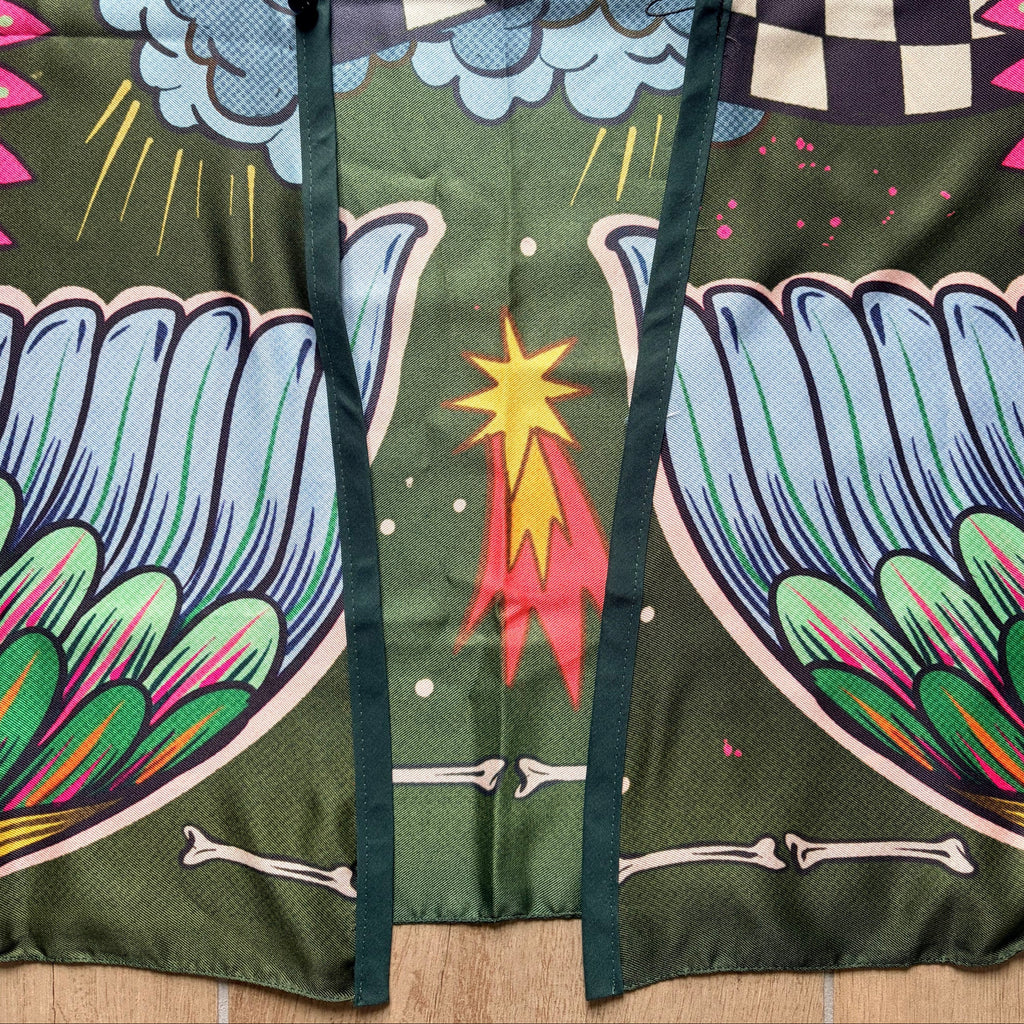 Dettaglio chiusura kimono "Prima o Poi" ARYK OLD PAINT – bottone nero su illustrazione con ali colorate, scacchi e stella cometa, fondo verde scur