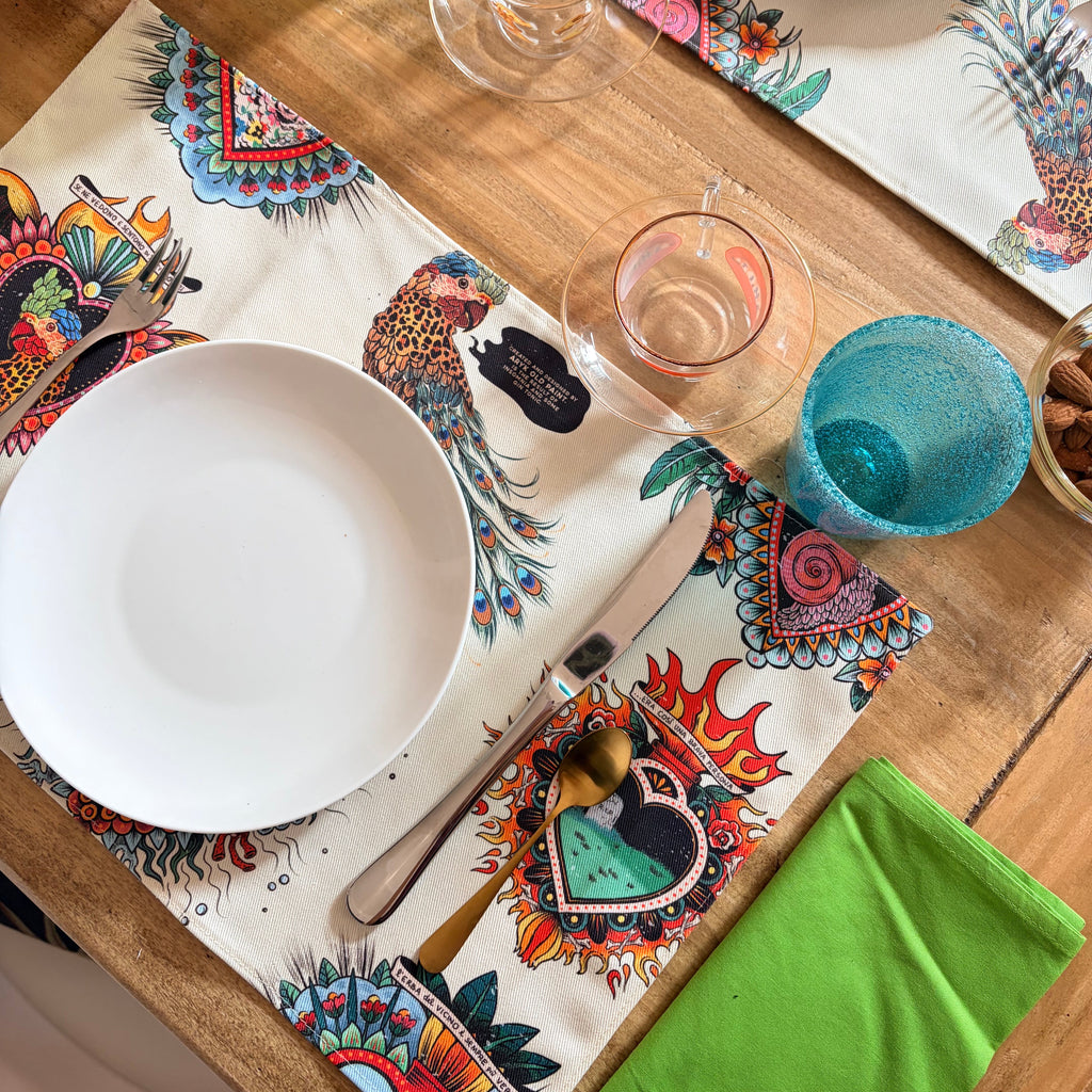  Set da 2 tovagliette "La Solita Zuppa" ARYK OLD PAINT su tavola apparecchiata con piatti bianchi e bicchieri – illustrazione old school con pappagallo leopardato e cuori sacri

