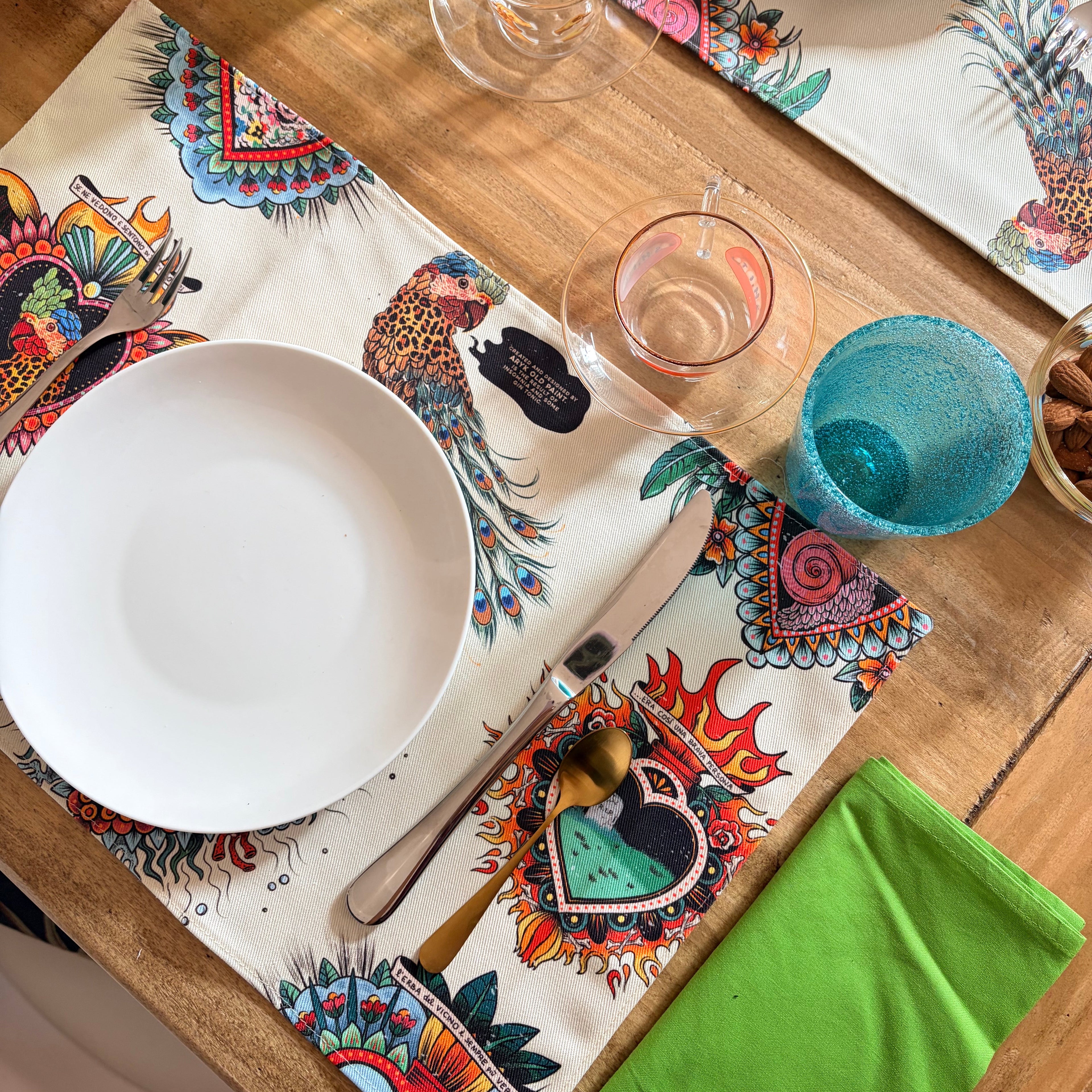  Set da 2 tovagliette "La Solita Zuppa" ARYK OLD PAINT su tavola apparecchiata con piatti bianchi e bicchieri – illustrazione old school con pappagallo leopardato e cuori sacri
